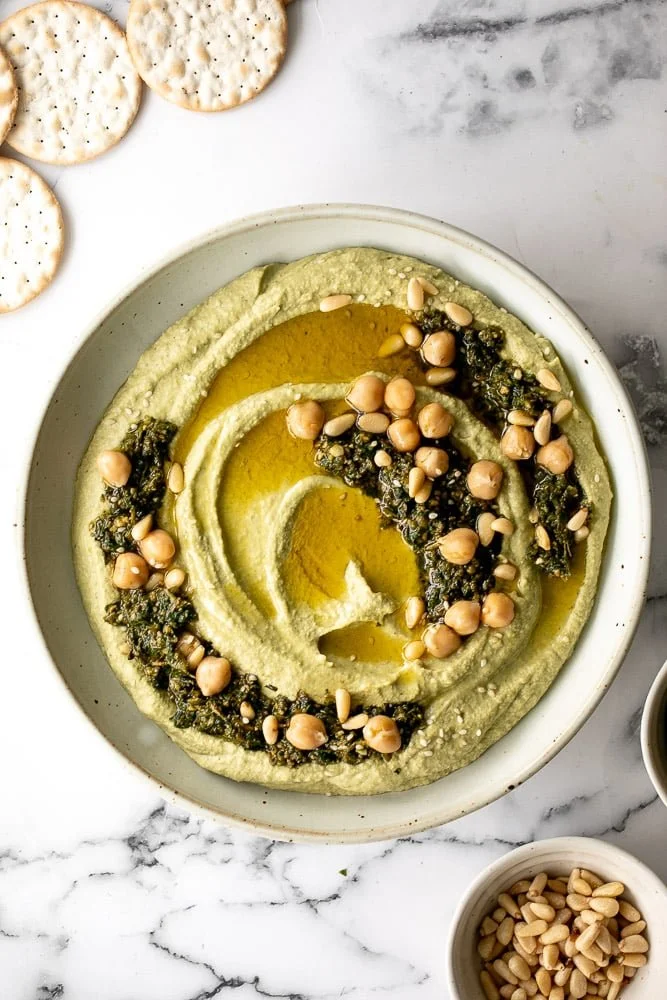A bowl of vibrant pesto hummus garnished with fresh basil and served with pita chips.